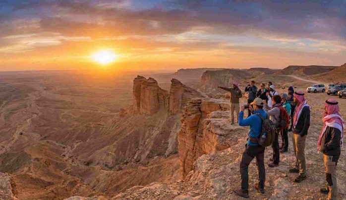 A group of tourists and a guide stand at the cliff's edge, enjoying the panoramic sunset view of the desert valley, a typical experience offered by the Best Edge of the World Riyadh Tours & Packages.