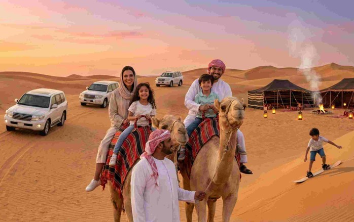 A smiling family enjoys camel rides and sandboarding at sunset in the dunes, with 4x4 vehicles and a traditional tent camp in the background, illustrating the experience of the Best Evening Desert Safari in Riyadh for Families.