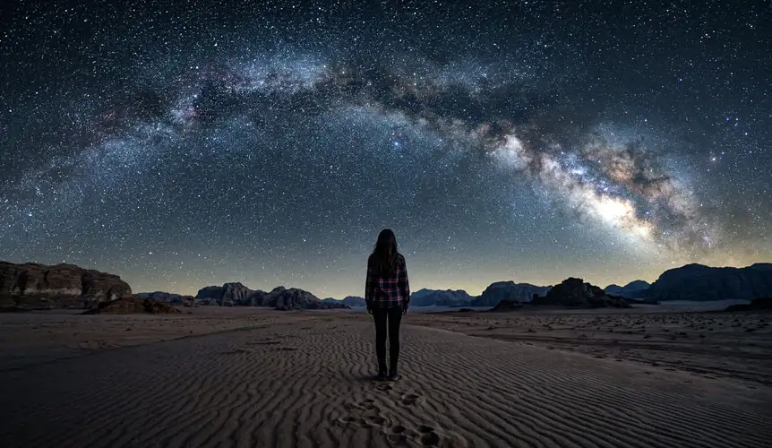 A person standing in the vast, dark sands of Saudi Arabia looking up at a brilliant starry sky, representing the humbling moments found in A Complete Guide to Stargazing in the Riyadh Desert.