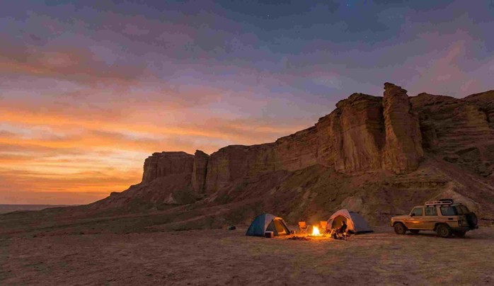 A scenic desert campsite with tents and a campfire near the dramatic cliffs at sunset, showcasing overnight stay possibilities featured in the Edge of the World Riyadh Hiking Guide.