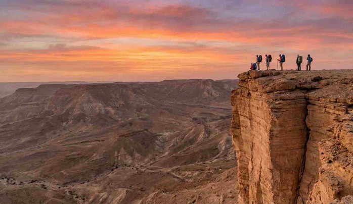 A group of hikers with backpacks standing on the sheer cliff edge at sunset, looking out over the vast desert canyon, illustrating a key destination featured in the Edge of the World Riyadh Hiking Guide.