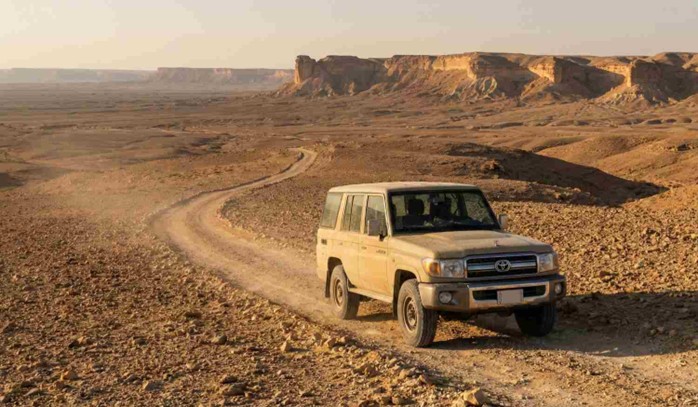 A rugged 4x4 vehicle driving on a winding desert track towards the distant cliffs, navigating the terrain to reach the location for Sunset at Edge of the World Riyadh (Photography Spots).