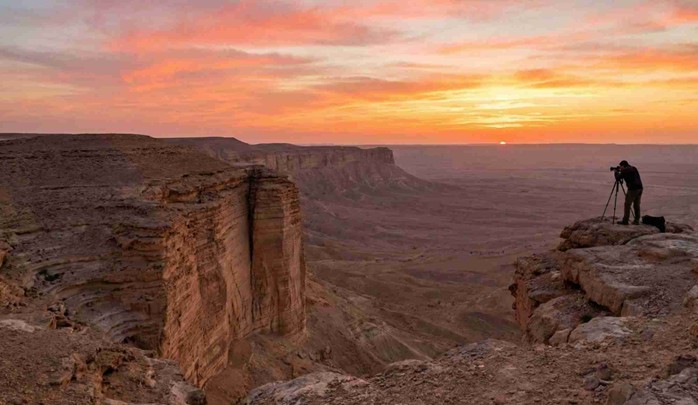 A photographer with a tripod stands on a dramatic cliff edge, capturing the stunning Sunset at Edge of the World Riyadh (Photography Spots) as the sun dips below the horizon, casting orange and pink hues over the vast desert valley and escarpment.