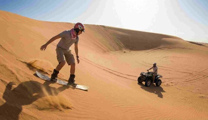 A man sandboarding down a large sand dune and another person riding a green ATV in the desert, showcasing some of the popular Evening Desert Safari Riyadh Activities (Dune Bashing, Camel Ride).