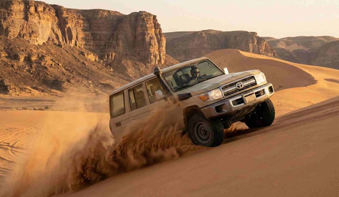 A beige Toyota Land Cruiser 4x4 vehicle is shown dune bashing, kicking up a large cloud of sand as it climbs a dune towards rugged cliffs at sunset, illustrating the type of off-road journey required for How to Get to Edge of the World from Riyadh.