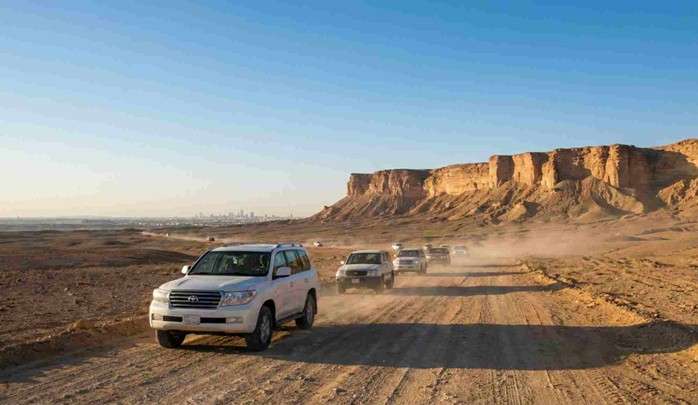 A convoy of 4x4 vehicles kicking up dust on a desert dirt road, heading towards a large escarpment with the Riyadh city skyline visible in the distance, illustrating the journey for How to Get to Edge of the World from Riyadh.