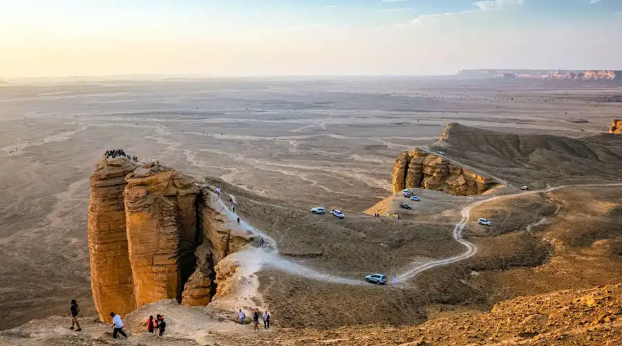 Rear view of a person in traditional dress riding a camel across sand dunes at sunset, with two 4x4 vehicles driving in the distance.