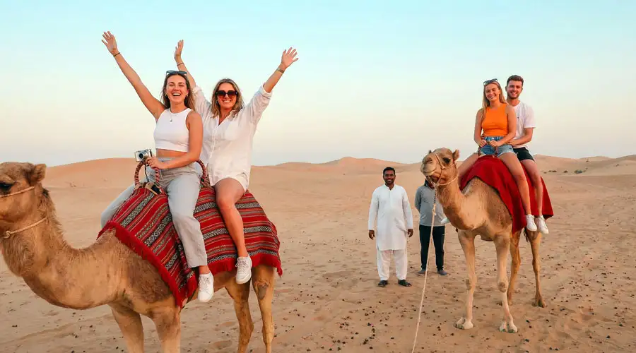 Two pairs of young tourists smiling and waving while riding camels led by a guide in a sandy desert in Riyadh.
