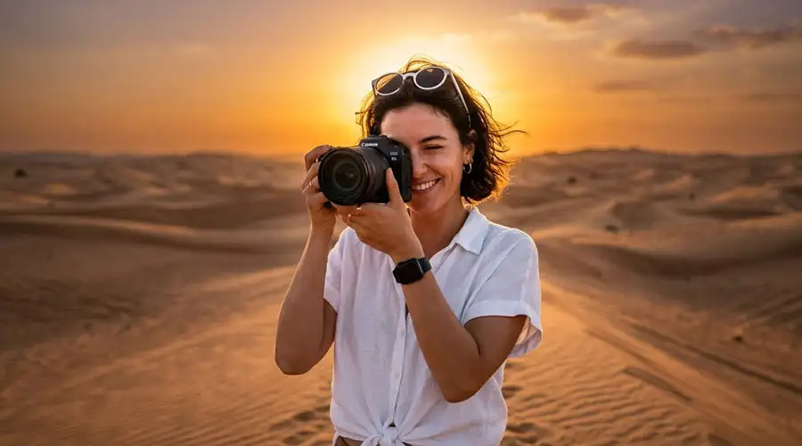 A smiling woman holding a professional DSLR camera to her eye, standing in front of rolling sand dunes during a golden sunset in Riyadh desert.